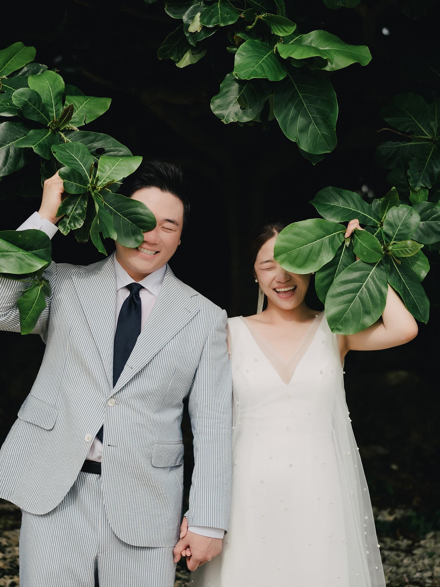 Bride and groom laughing behind big green leaves in Okinawa