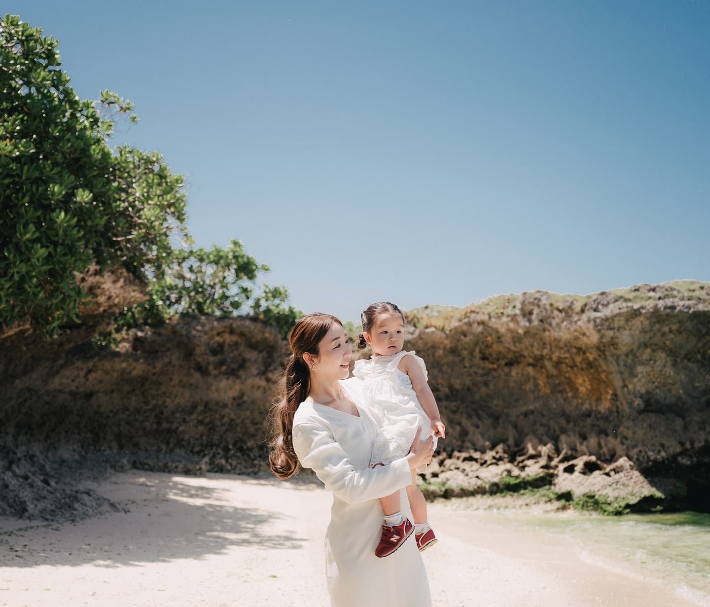 Mother carrying her baby daughter on a rock-lined Okinawa beach under a clear blue sky