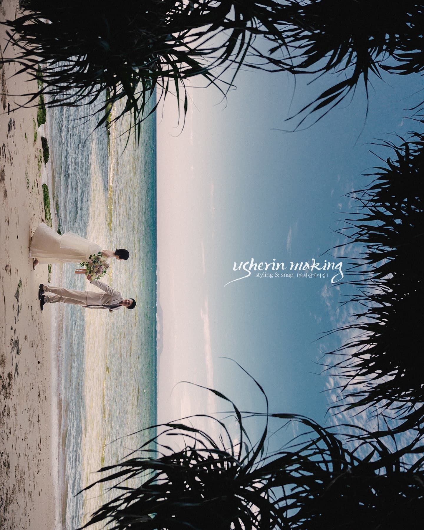 Bride and groom on a white sand beach framed by pandanus leaves, Okinawa's emerald sea behind