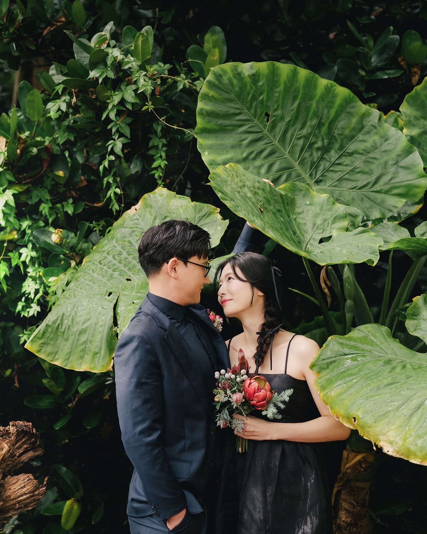 Couple among giant green leaves with a red bouquet in rainy Okinawa
