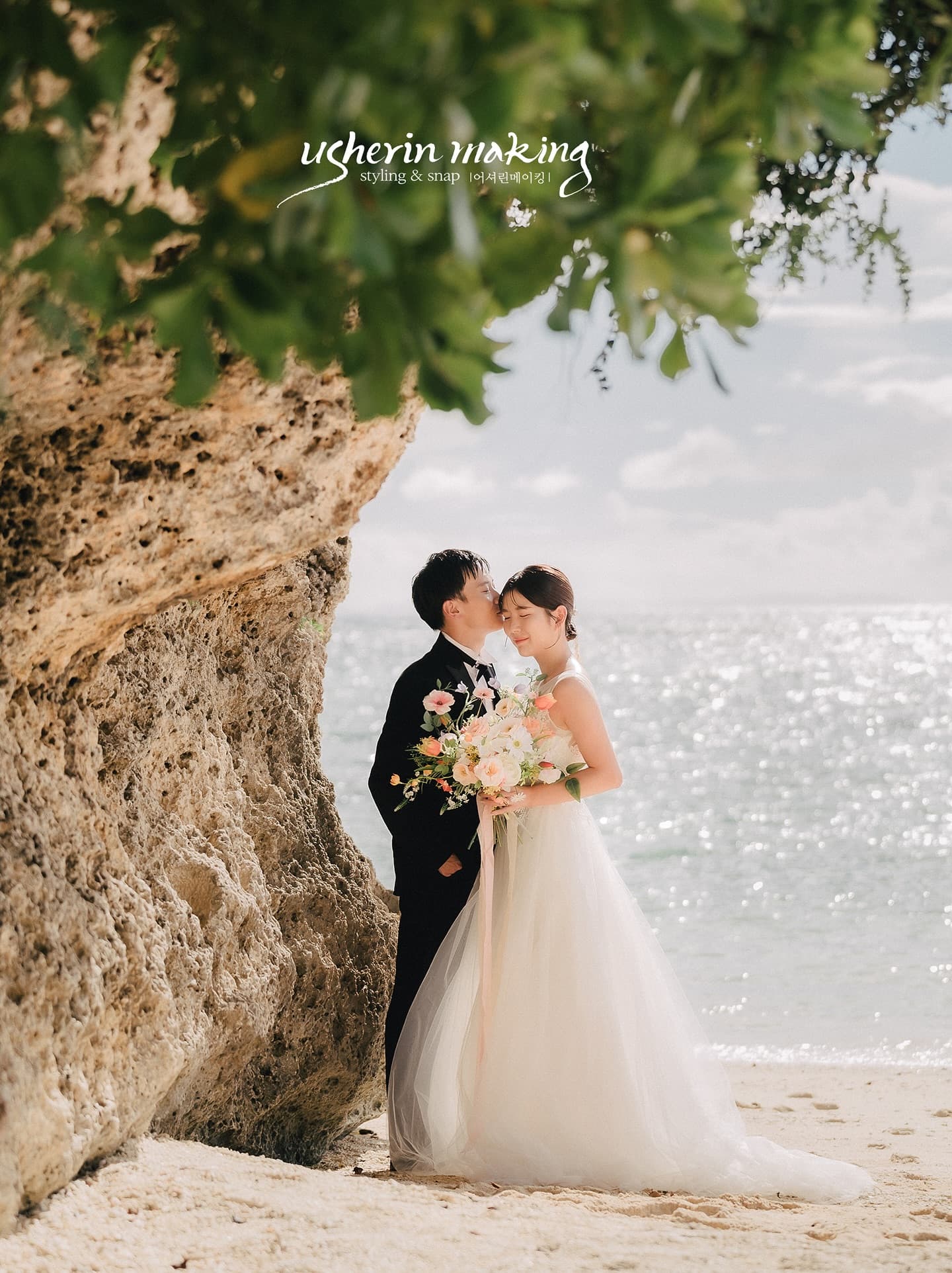 Groom kissing the bride's forehead beside a coastal rock with the bright sea behind in Okinawa