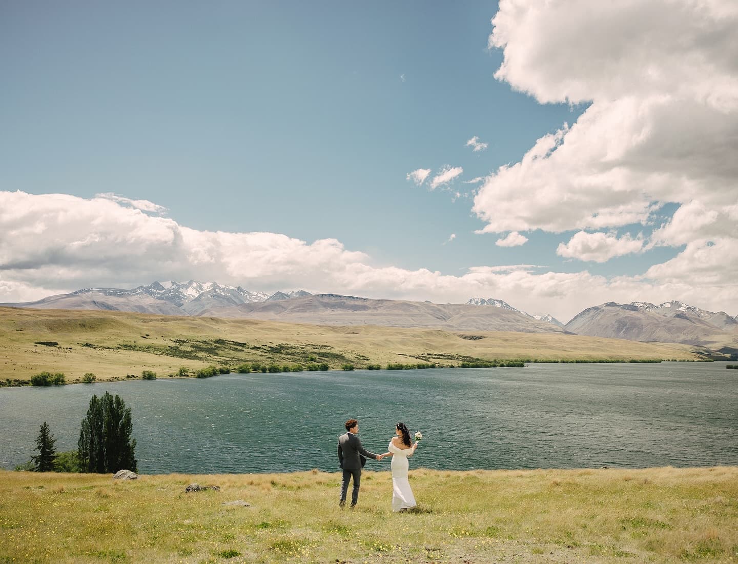 Bride and groom joining hands in a grassland before a lake and mountain range in New Zealand