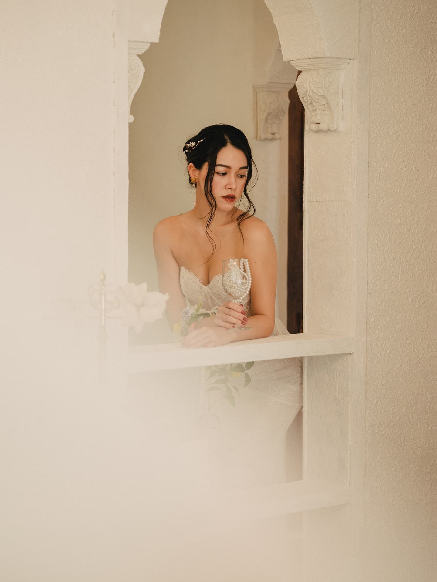 A bride lingers at an arched white window in a softly lit studio