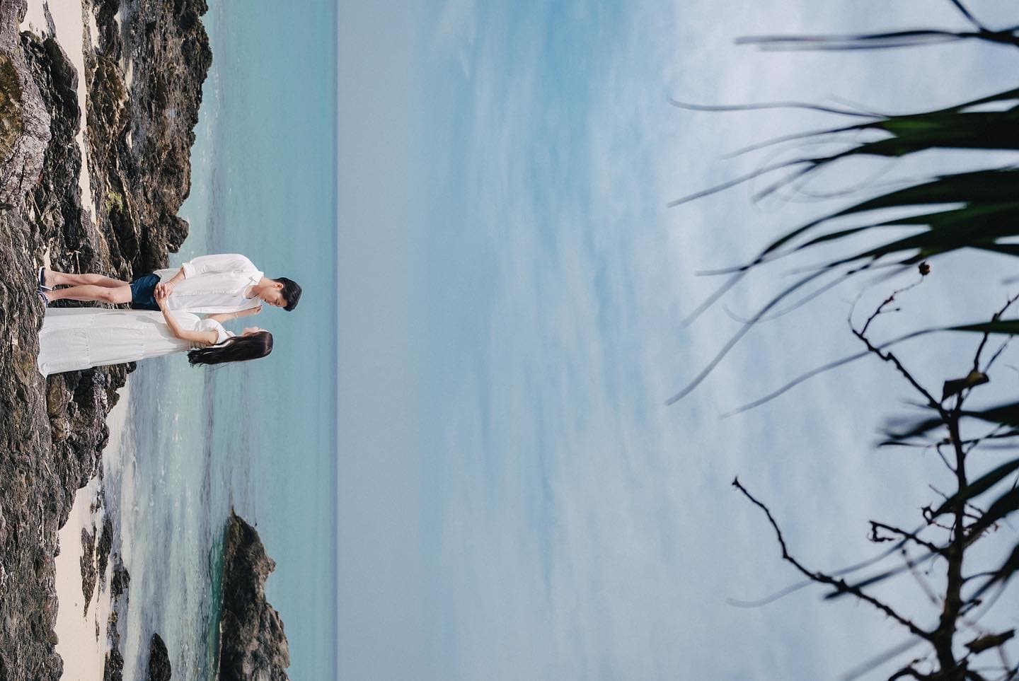 Couple standing close on shoreline rocks under a blue sky, Okinawa