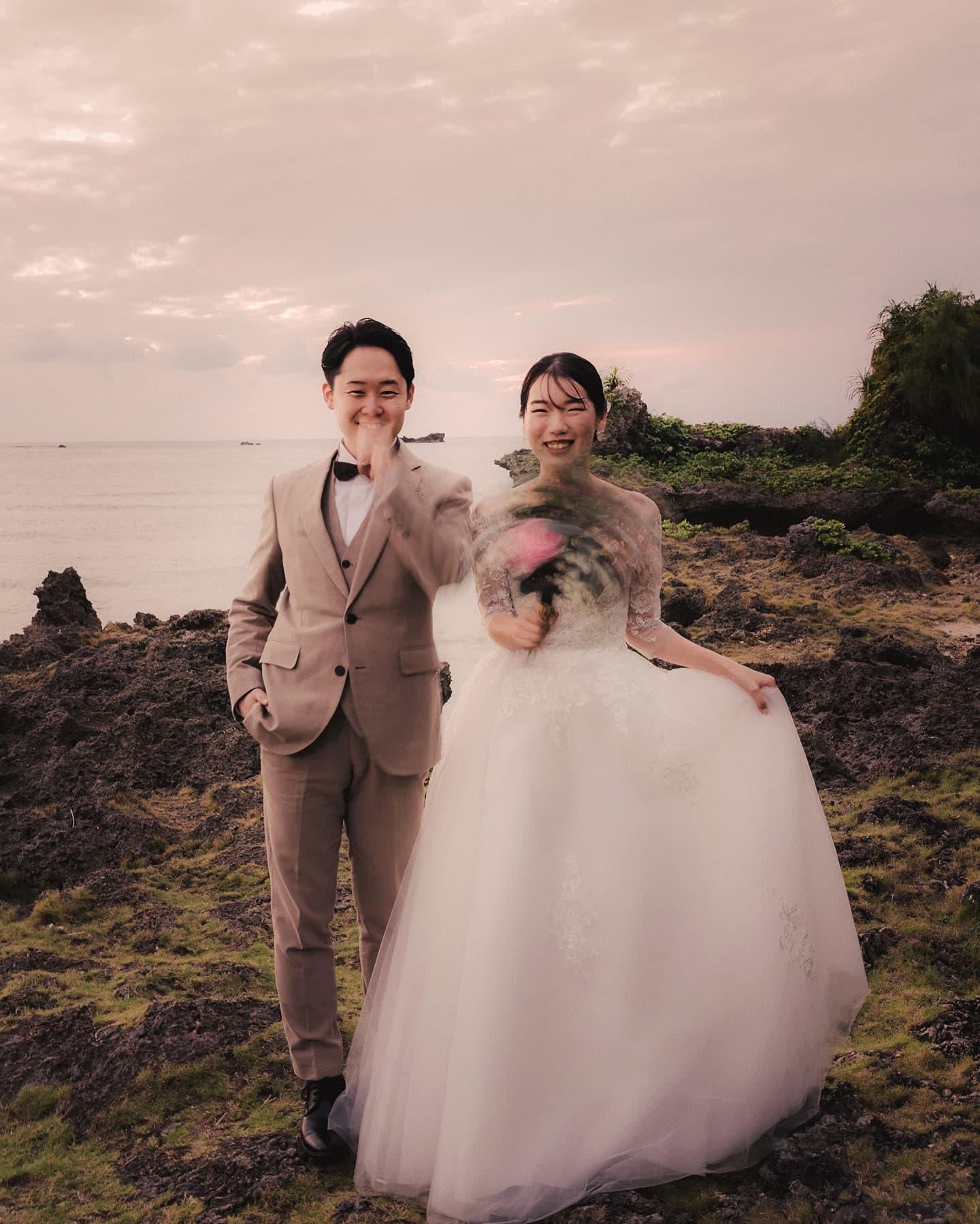 Couple laughing on a rocky Okinawa shore under a pale pink sunset sky