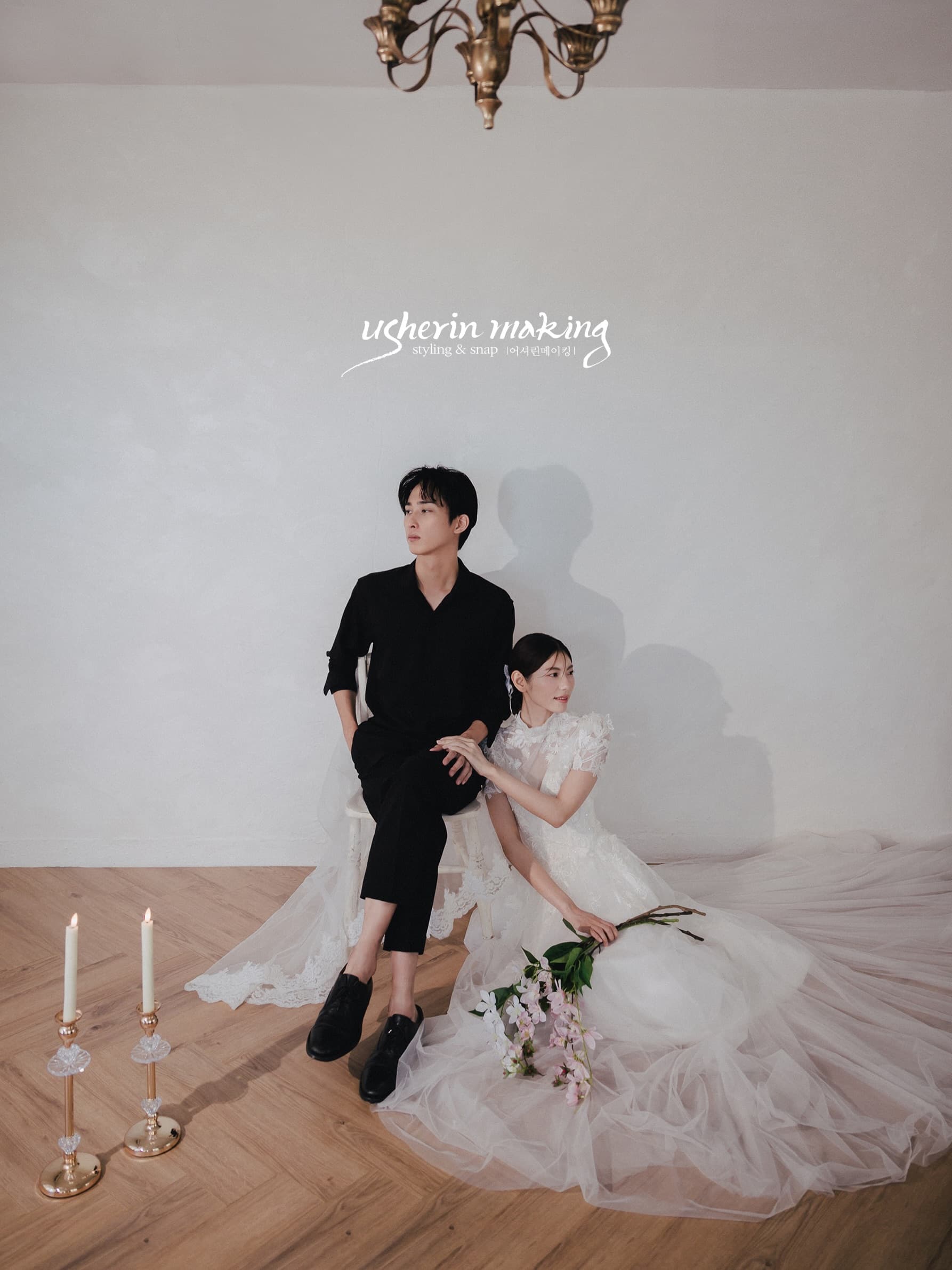 Groom in black seated beside the bride on the floor with a pink bouquet under a chandelier