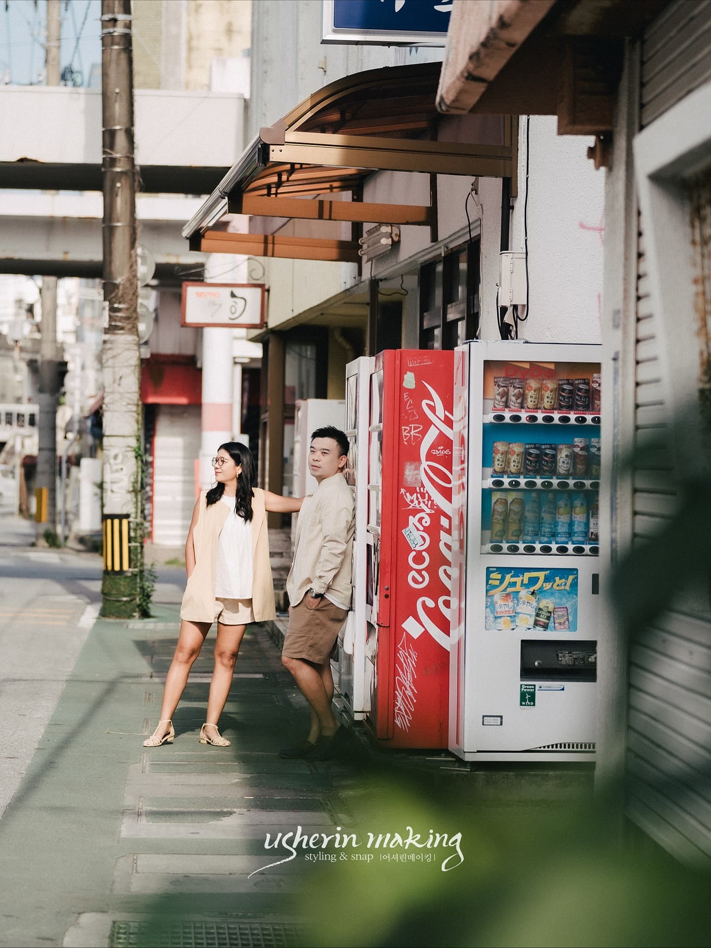 Casual couple posing by a red vending machine on a quiet Okinawa street