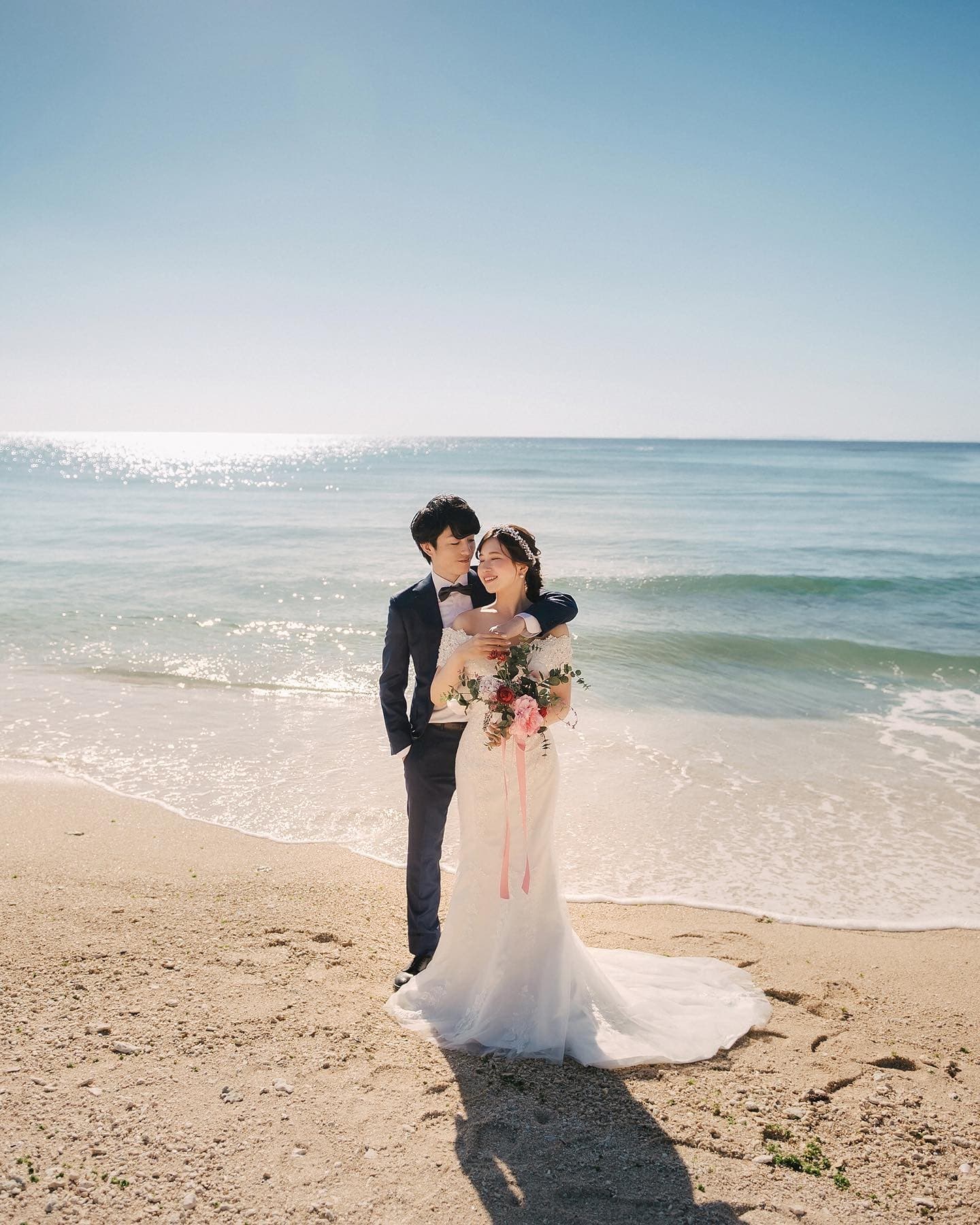 On a clear day, a groom embraces his bride from behind on an Okinawa beach