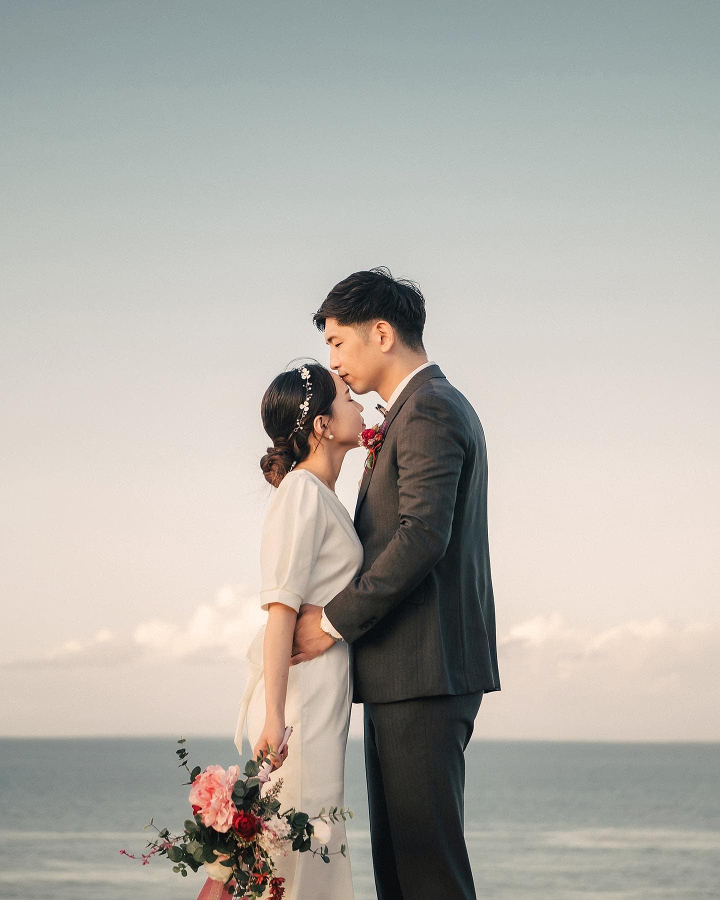 静かな海を背に、赤いブーケを持つ新婦の額にキスする新郎