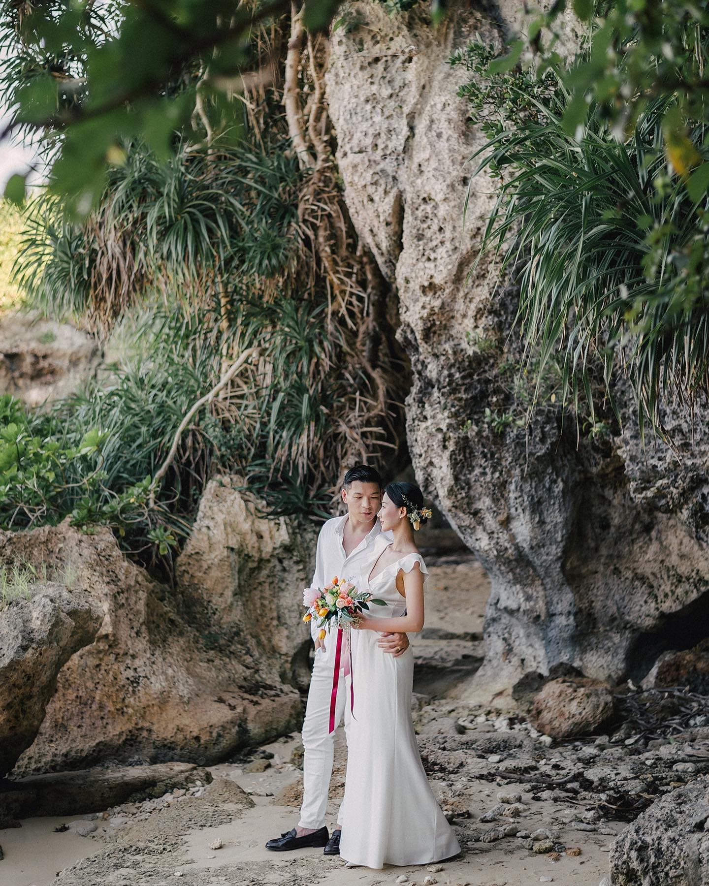 Couple in white holding a bouquet among rugged rocks draped with greenery in Okinawa