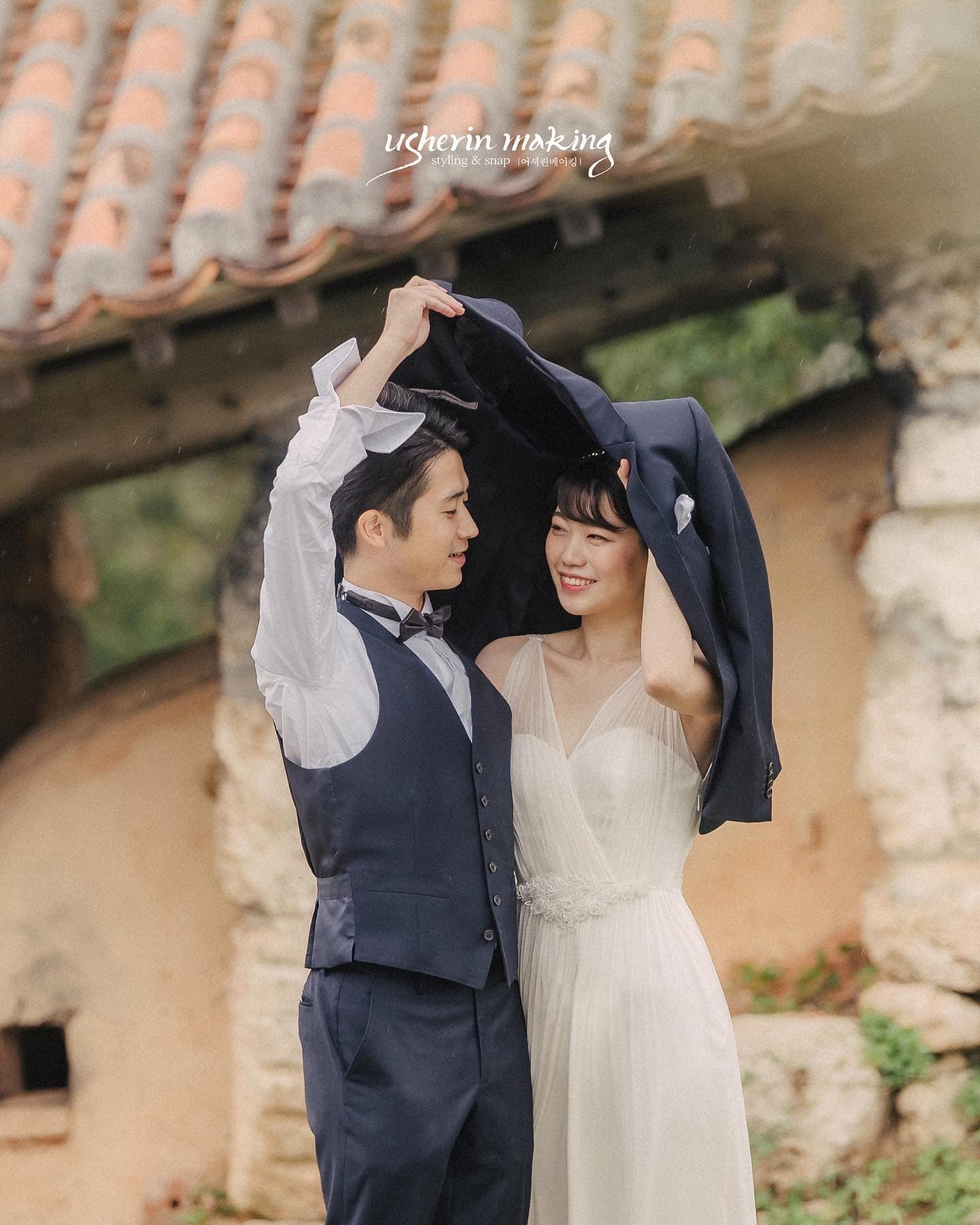 Bride and groom smiling under a jacket held like an umbrella by a red-tiled Okinawan house