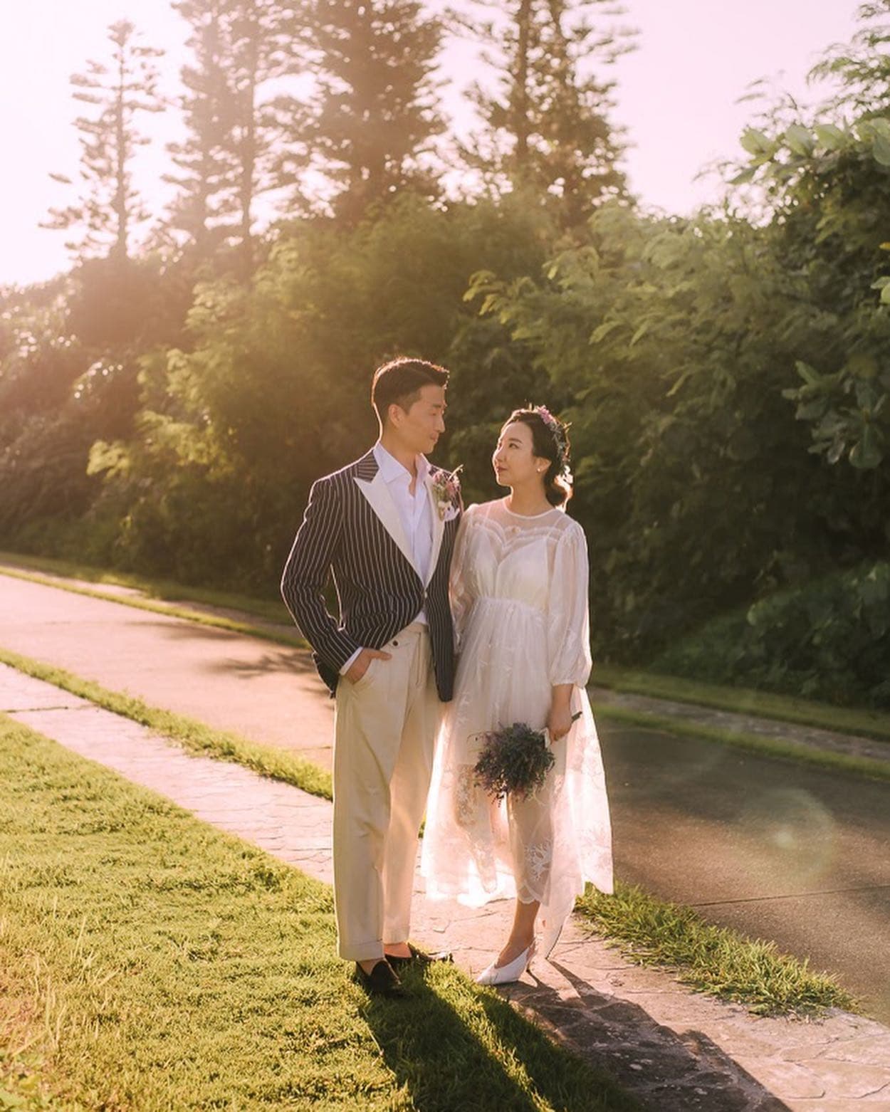 Couple on a tree-lined Okinawa path in golden backlight, bride in a sheer white dress