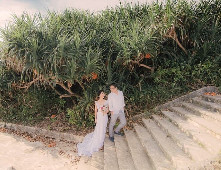 Bride and groom on stone steps by an Okinawa beach, lush pandanus trees behind