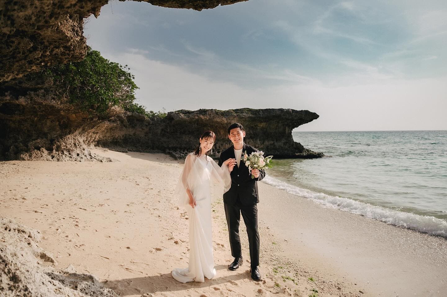 Newlyweds smiling beneath a rock overhang beside the sea in Okinawa