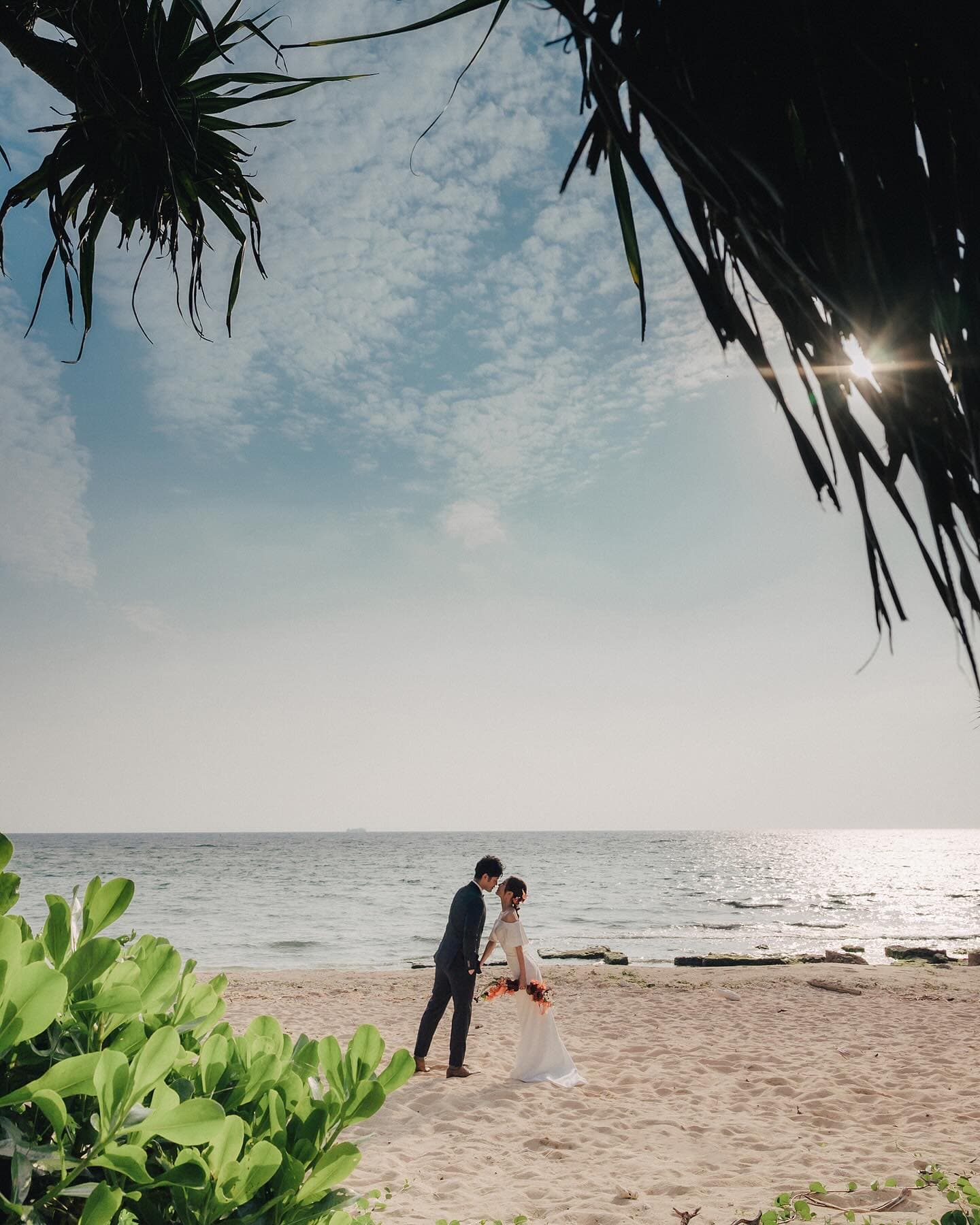 Couple kissing on a white sand beach framed by palm leaves under a summer sky in Okinawa