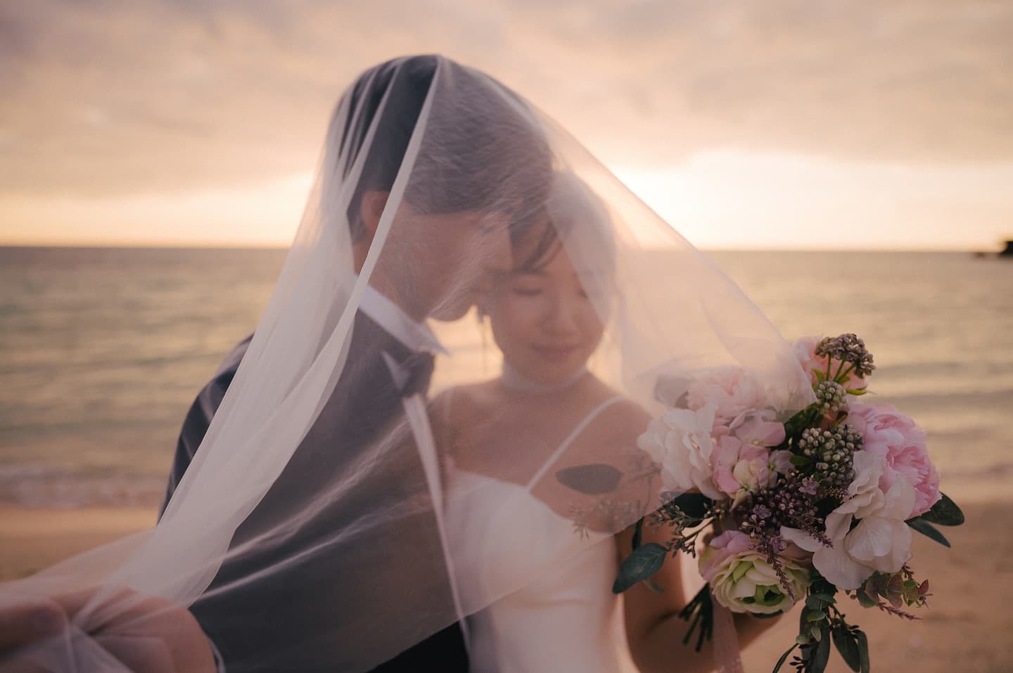 Couple close together under a sheer veil with a bouquet at sunset on an Okinawa beach